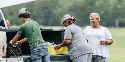 Three people unloading the back of a pickup truck in a rural area.
